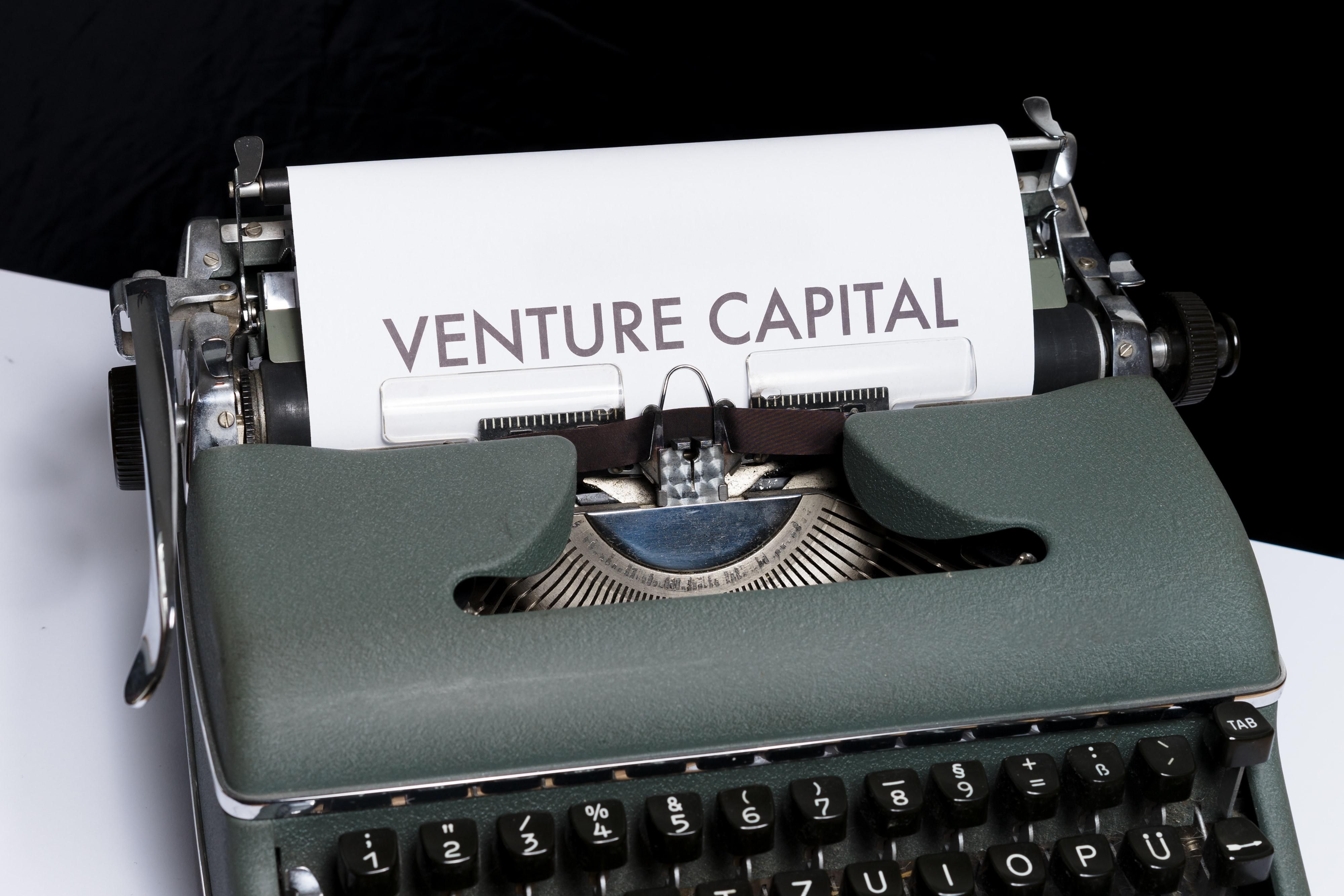 Egy szürkés-zöldes írógépen a Venture Capital szavakat gépelték be, ami kockázati tőkét jelent, az írógép egy fehér asztalon van, a háttér sötét