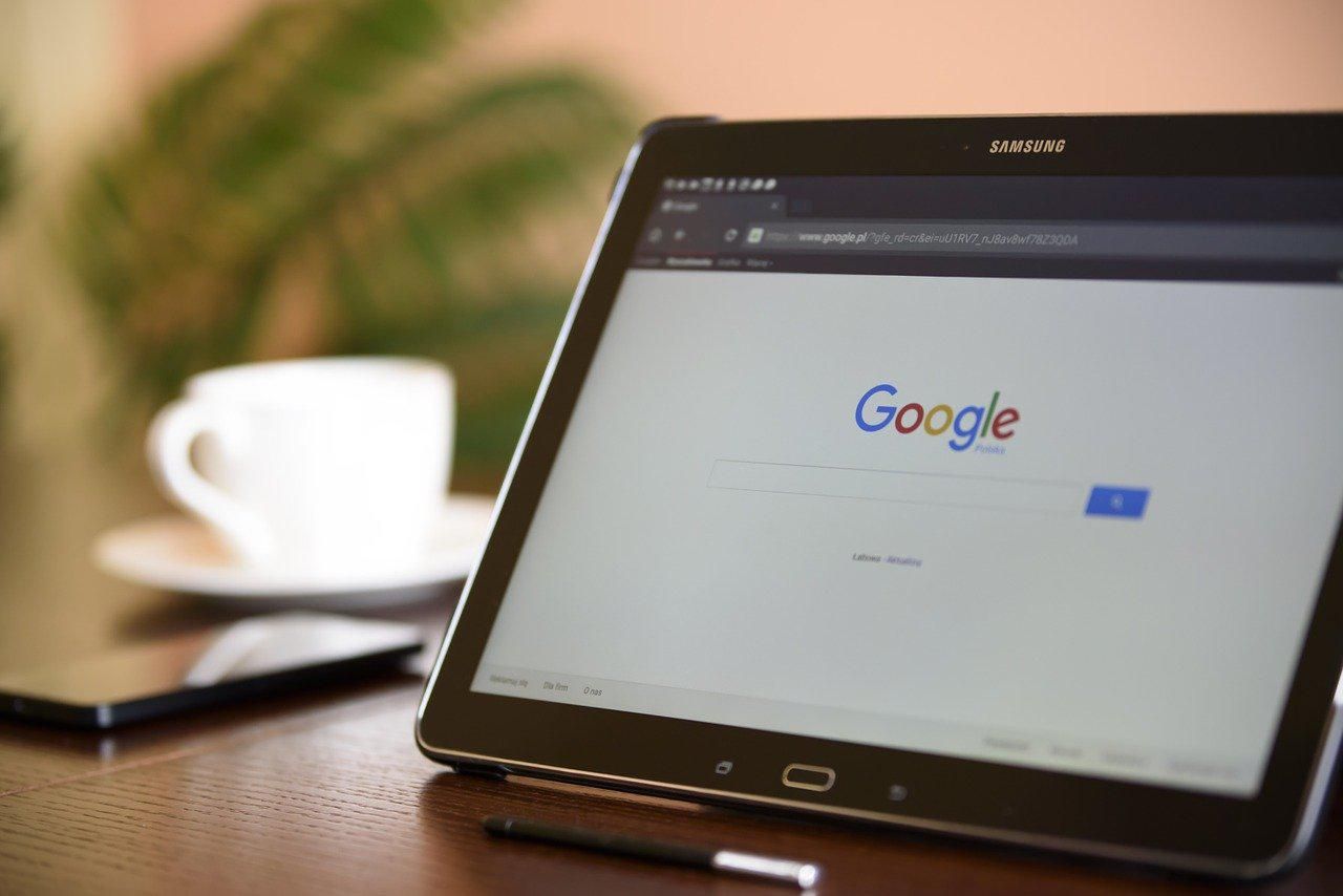 Egy fa asztalon egy kávéscsésze, egy okostelefon és egy toll mellett áll egy tablet, amelyen a Google nyitóoldala látható