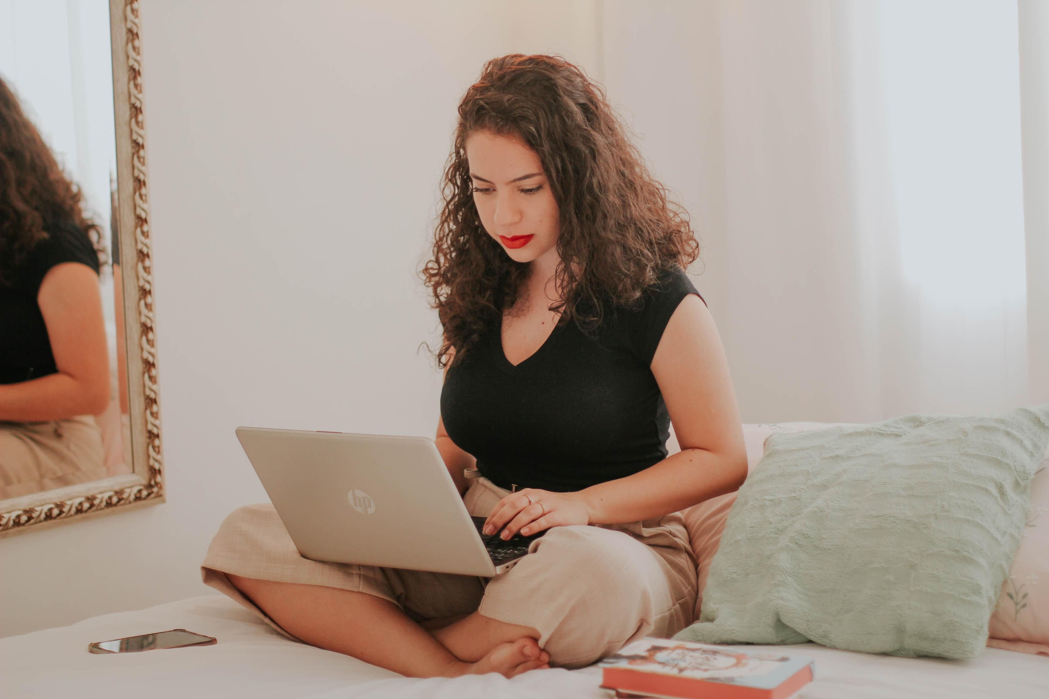 Dekoratív lány világos nadrágban, fekete felsőben ül az ágyán home officeban ölében egy laptoppal és dolgozik, mellette a falon egy tükör van