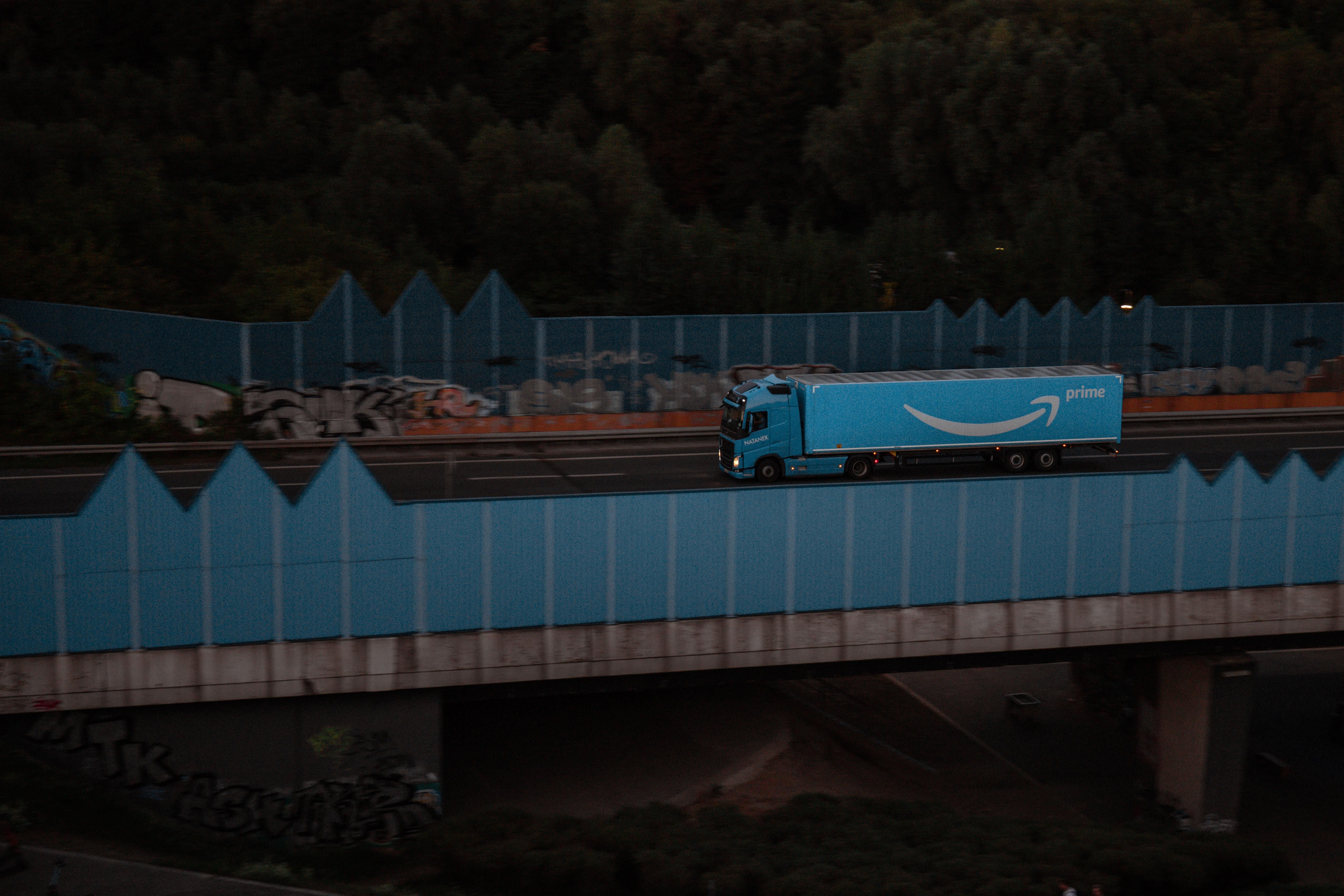 Egy kék színű Amazon Prime feliratos kamion halad át egy kék színű összefirkált fallal védett hídon esti szürkületben