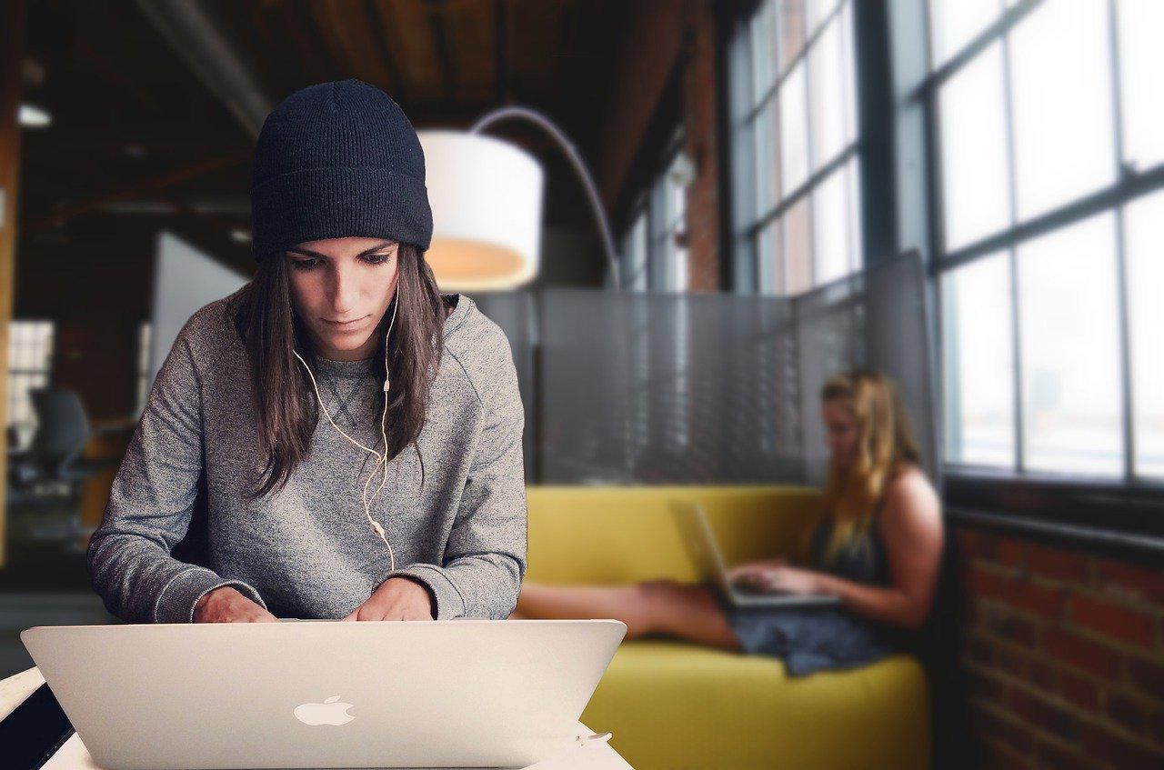 Sapkás, szürke pulcsis lány fehér fülhallgatóval dolgozik az Apple gépén egy másik nő előtt, aki egy sárga kanapén ül