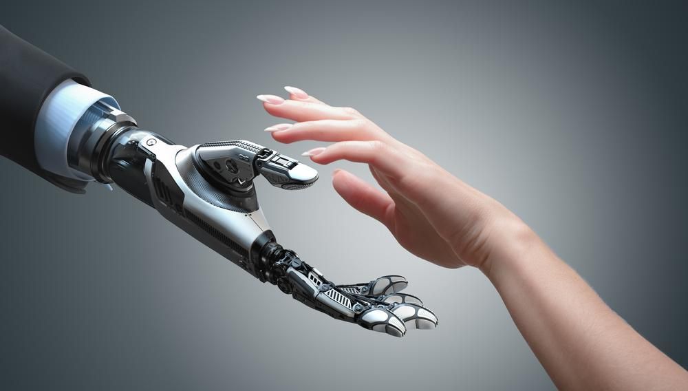 Mesterséges intelligenciával felvértezett robot nyújtja a kezét egy kifestett körmű nőnek egy szürke szobában