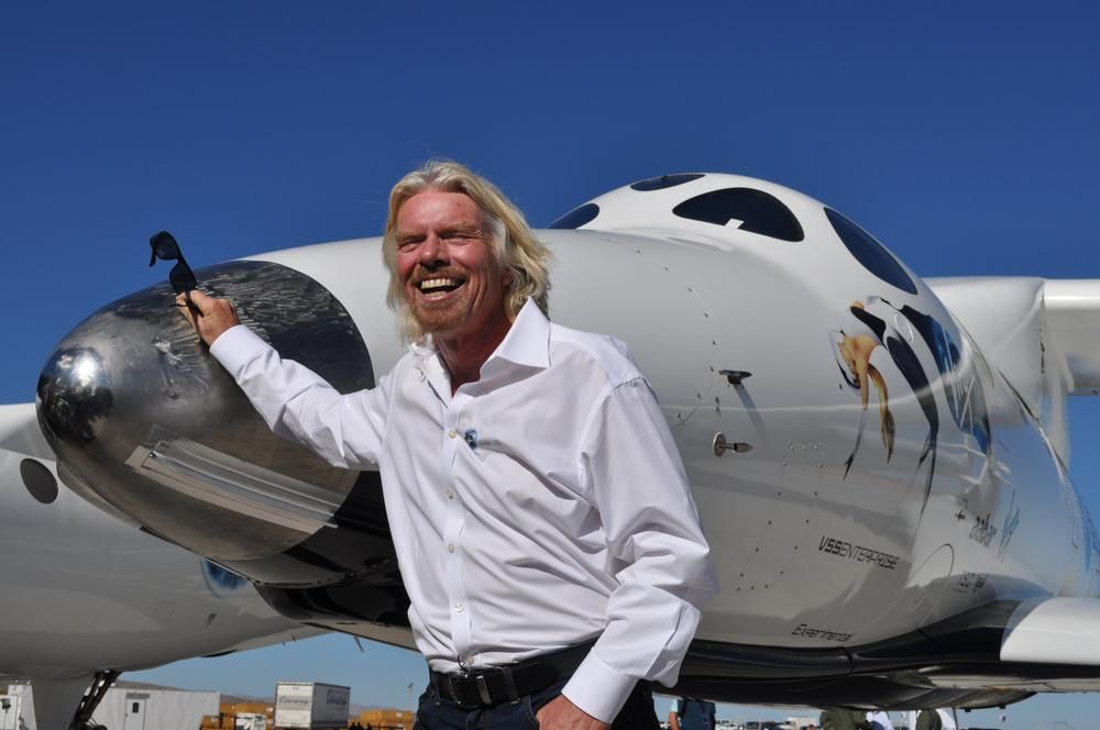Sir Richard Branson fehér ingben nevet a VSS Enterprise űrhajó előtt