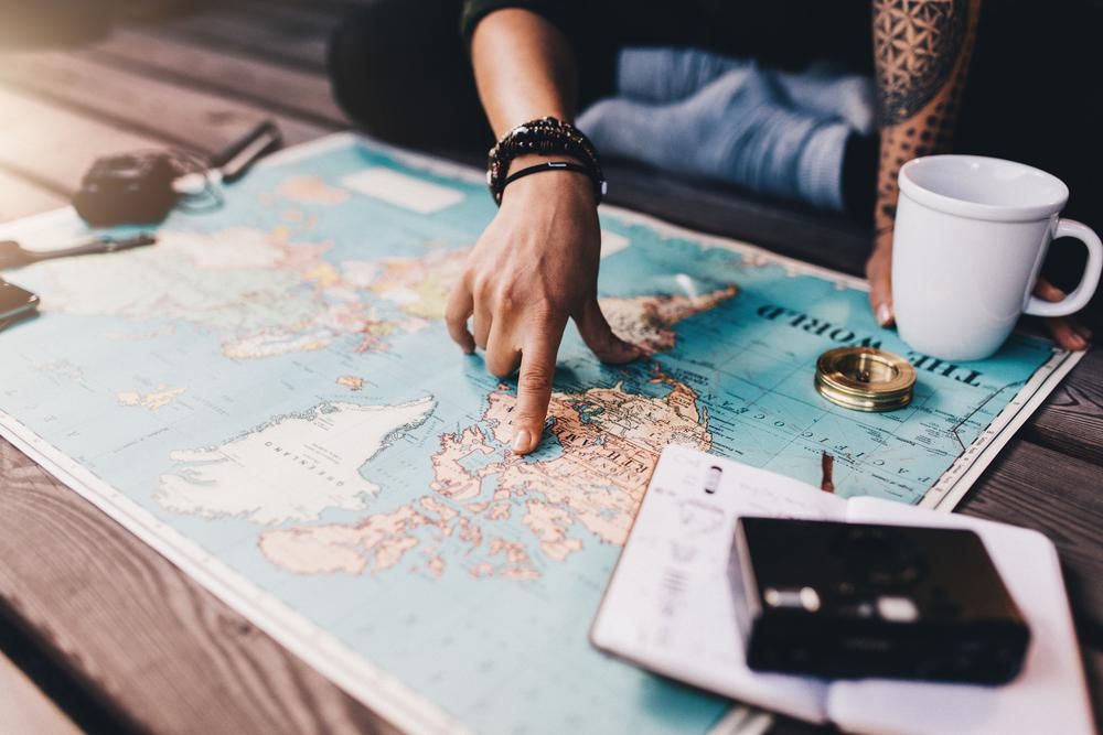 Tetovált ember a Föld térképén nézelődik és rámutat egy pontra, a térkép egy fa asztalon van, rajta egy bögre és egy iránytű