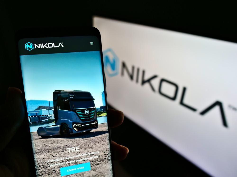 A Nikola elektromos kamiongyártó vállalat applikációja egy telefonon, amit egy ember tart a kezében, a kijelzőn egy kék kamion látható, a kép hátterében elmosódottan a Nikola logója látható