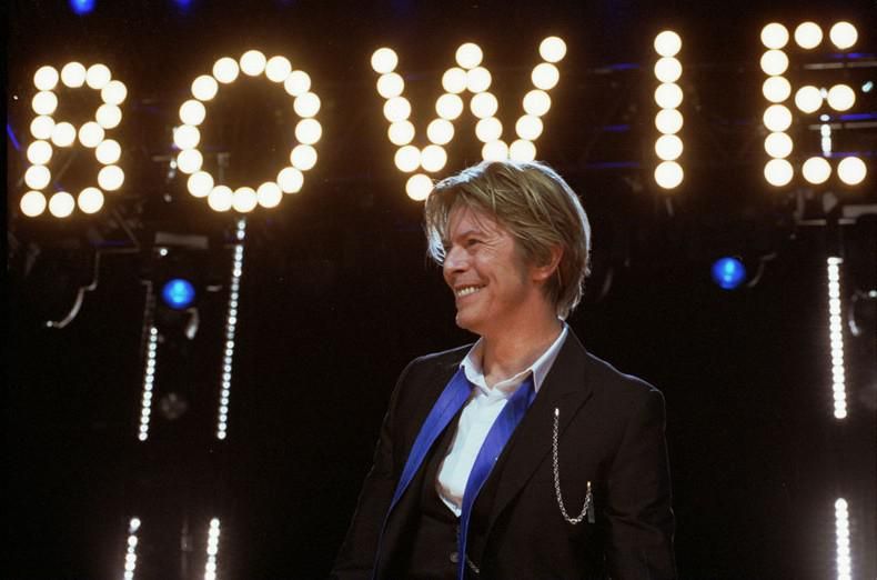 David Bowie a színpadon fekete zakóban, szőke hajjal, egy világító Bowie felirat alatt nevet