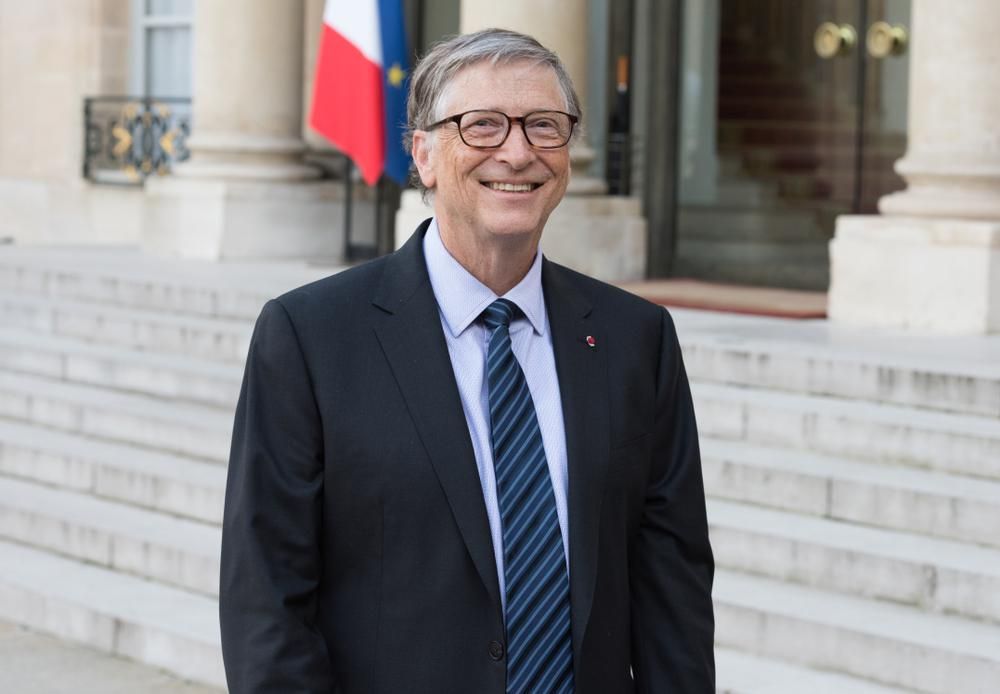 Bill Gates fekete zakóban, kék ingben és csíkos nyakkendőben vigyorog egy épület lépcsője előtt