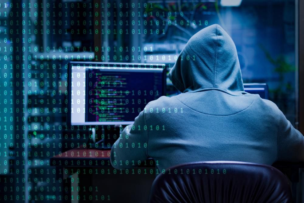 Fehér kapucnis pulcsi hacker ül egy számítógépnél, miután ingatlanportálokat fertőzött meg, a képen bináris számsorozat látható