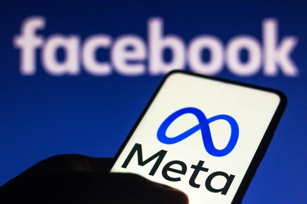 A Meta logója egy okostelefonon, ami egy ember kezében van, a háttérben a Facebook felirata látható egy kék felületen