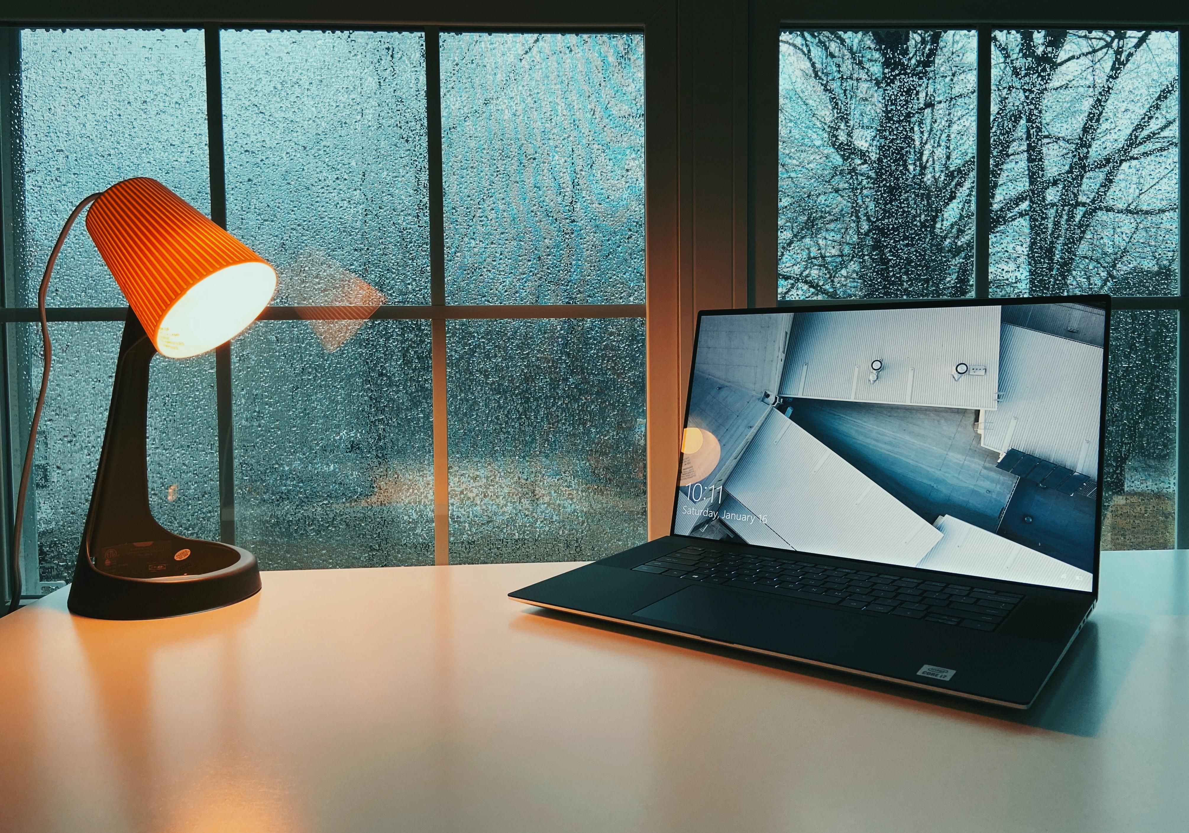 Egy narancssárga búrájú, fekete vázú asztali lámpa megvilágít egy fehér asztalt, amin egy laptop/PC található, az asztal egy ablak előtt van, kint esik az eső