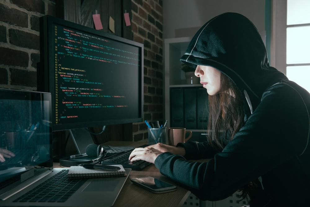 Egy kapucnis nő éppen behackeli magát egy rendszerbe, hogy letitkosítsa az adatokat és váltságíjat követeljen, egy telefon és egy másik laptop is látható a képen