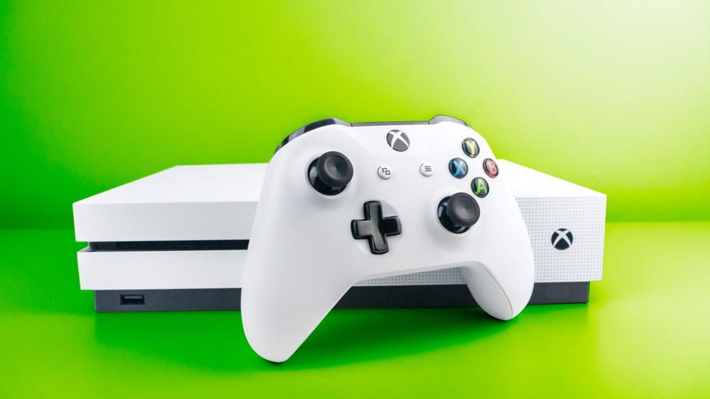 Fehér színű Xbox One konzol és a hozzá tartozó joystick egy neonzöld környezetben