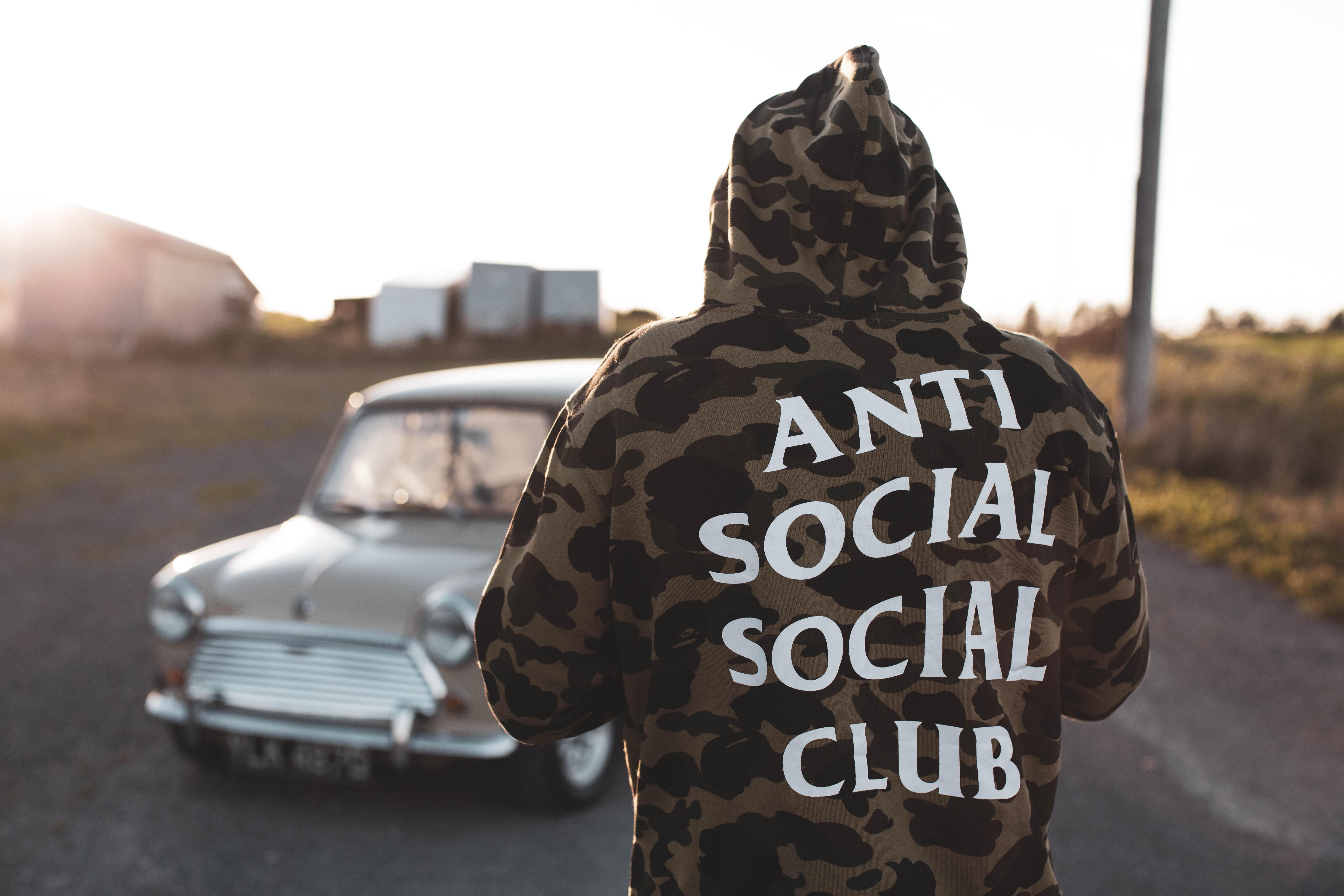 Egy terepmintás pulóveres férfi háttal áll a kamerának, nézi az autóját, kapucni van a fején, a pulcsin antiszociális közösségi klub felirat látható