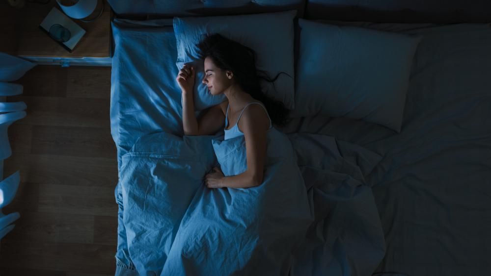Egy fekete hajú nő alszik a fehéres színű pizsamájában és ágyneműjében, be van takarózva, laminált parketta és félhomály van a szobában