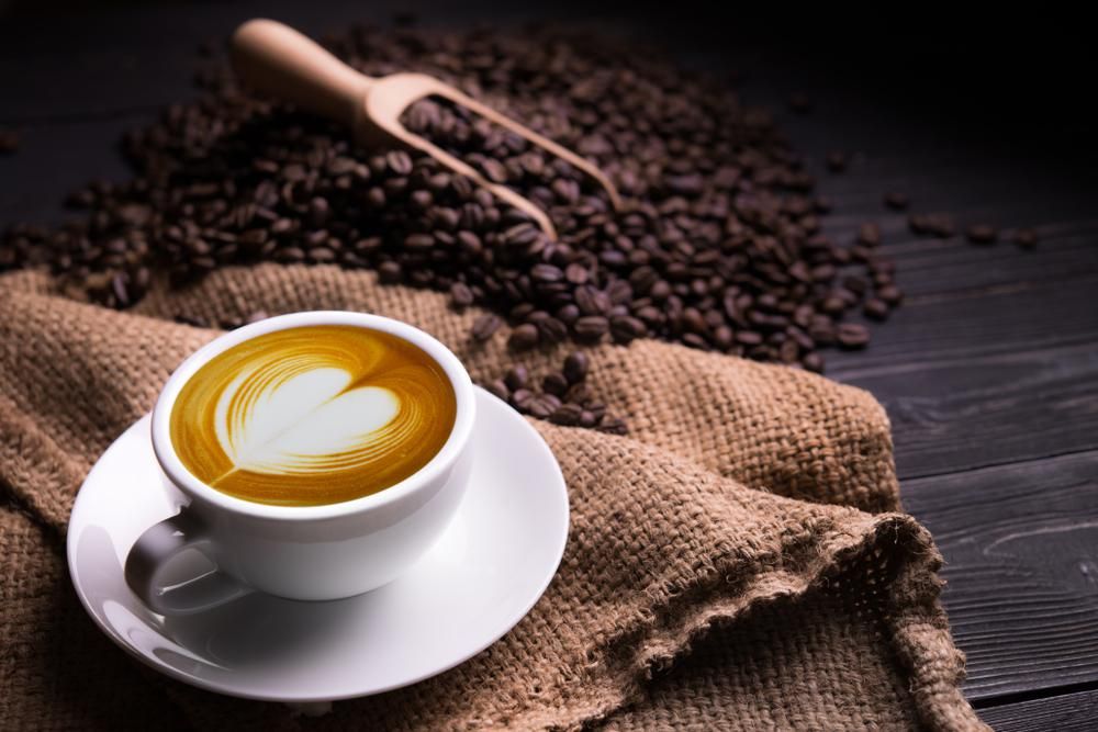 Kávébabok és egy szív alakú habbal elkészített csésze kávé egy zsákon, ami egy fa felületen helyezkedik el