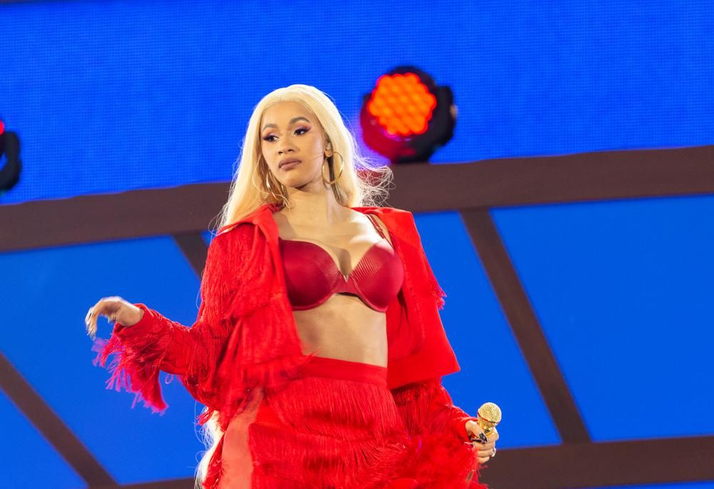 Cardi B amerikai rapper piros ruhában, piros melltartóban, szőke hajjal egy színpadon áll, épp rapkoncertje van, kezében egy arany mikrofont fog