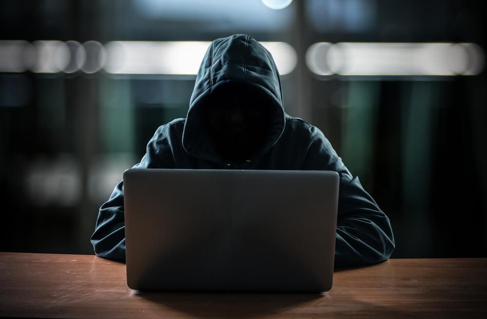 Kapucnis hacker ül a gépe előtt, ami egy fa asztalon van