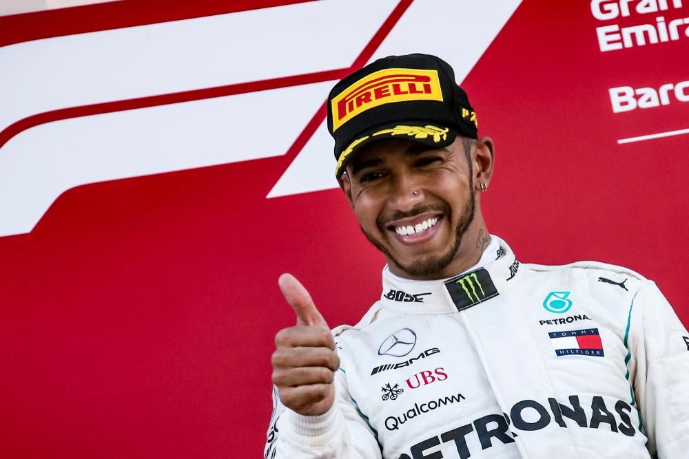 Lewis Hamilton a hüvelykujját mutogatja fehér versenyruhájában egy piros-fehér háttér előtt, miközben vigyorog