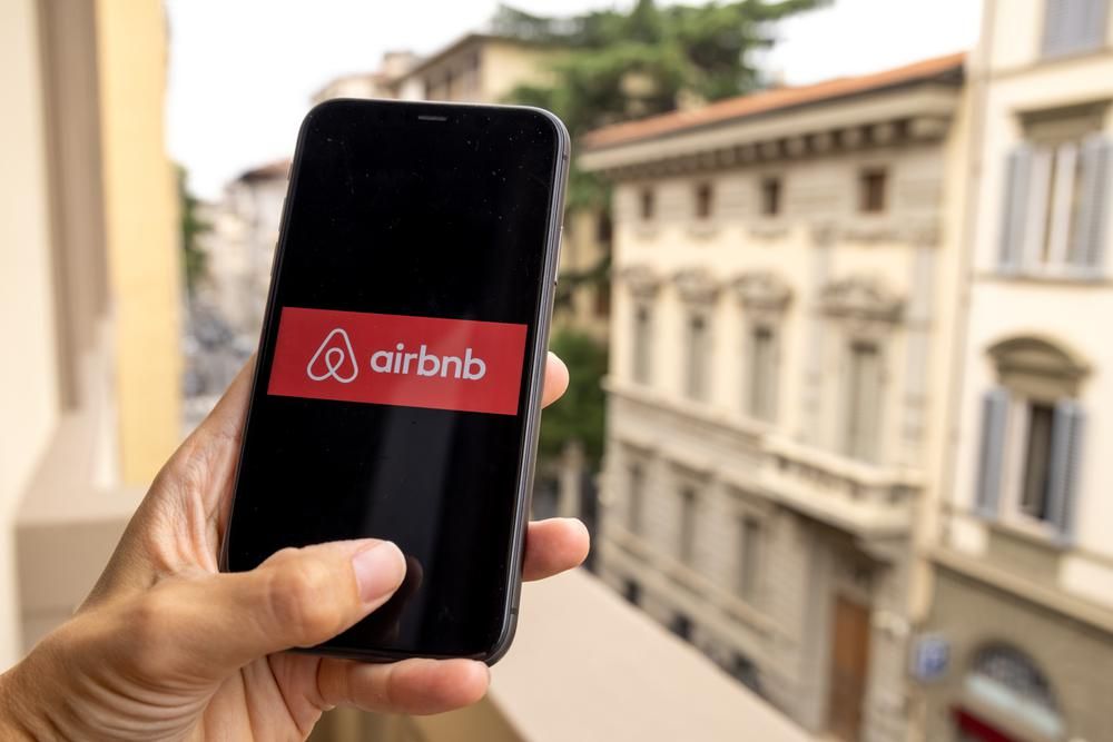 Egy nő kezében tartja okostelefonját, a képernyőn az Airbnb logója látható, a háttérben egy olasz település van