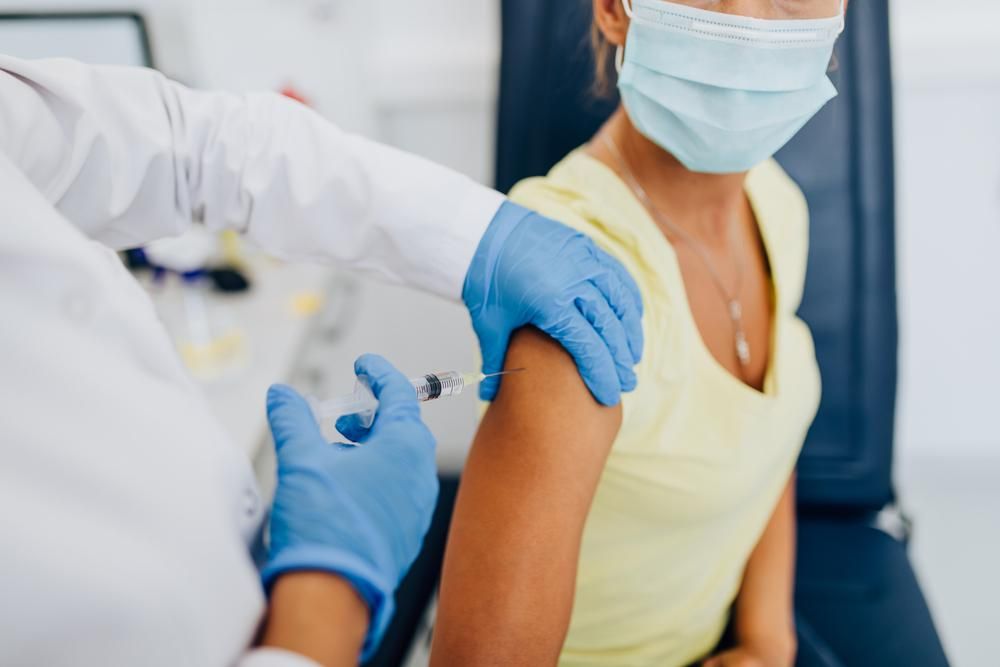 Egy ápolónő épp koronavírus elleni vakcinát ad be egy sárga felsős, maszkos hölgynek