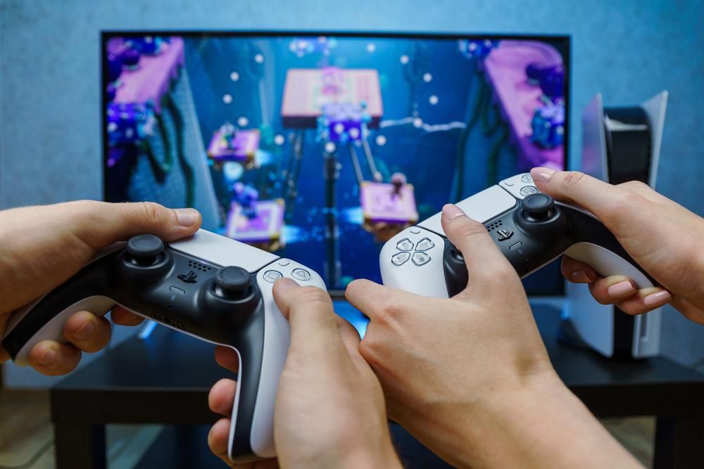 Egy férfi és egy nő a Sony Playstationjén játszik, fekete-fehér kontrollerekkel, kék fal előtt