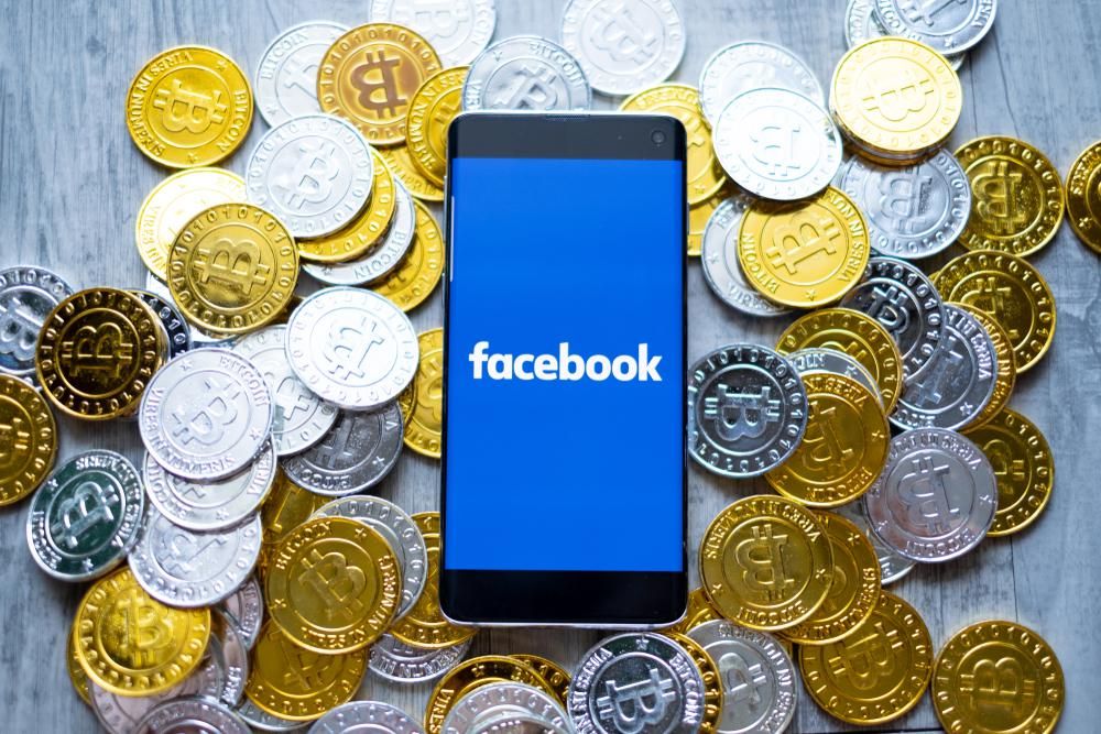 Egy okostelefon, amelynek képernyőjén a Facebook logója látható, a telefon körül kriptovaluta, pontosabban bitcoin fantáziaérmék láthatók