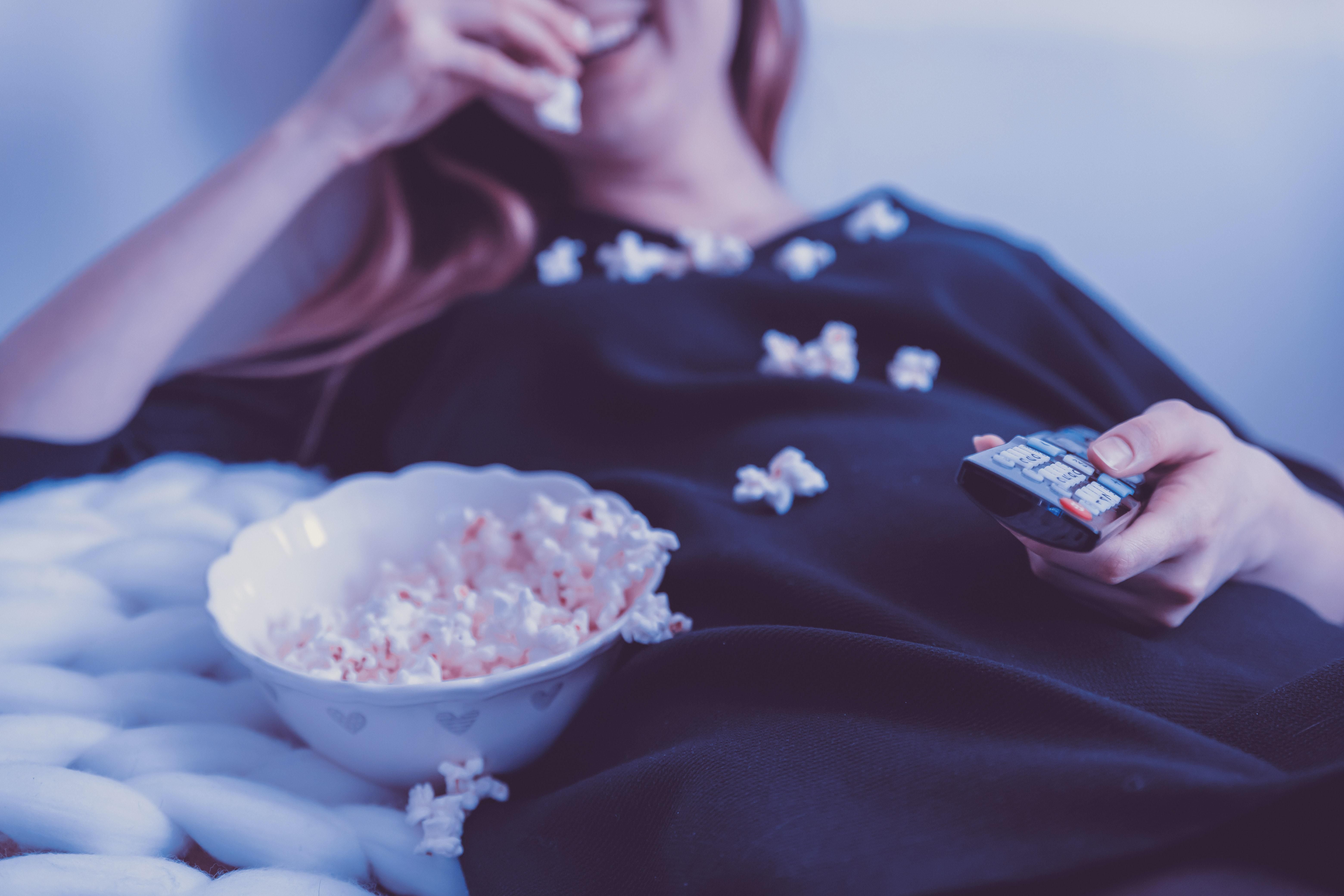Fiatal lány nevet, miközben TV-t néz egy sötétkék felsőben, közben popcornt eszik