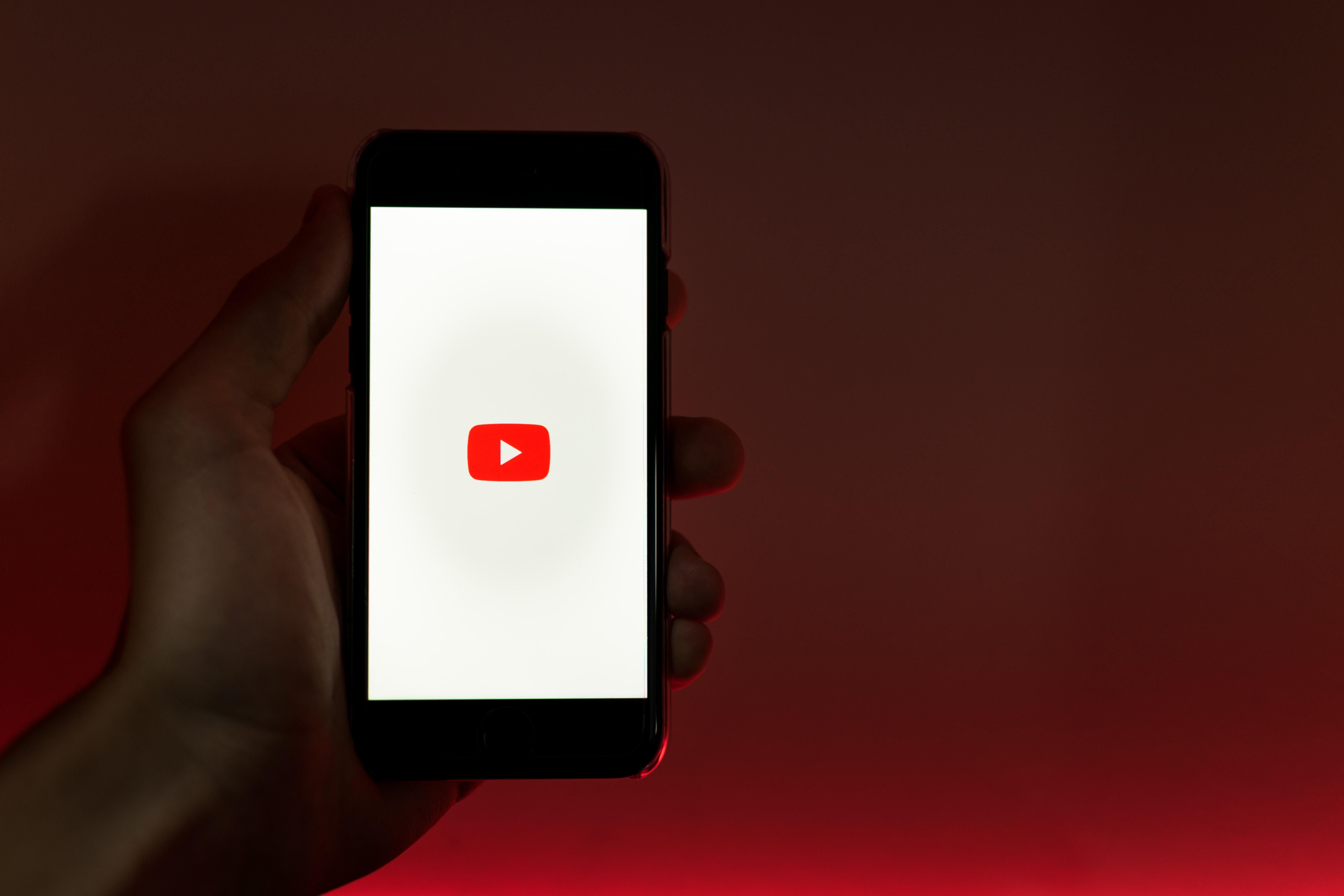 Egy ember egy okostelefont tart a kezében,a melynek képernyőjén a YouTube logója látható. A háttér piros-fekete.