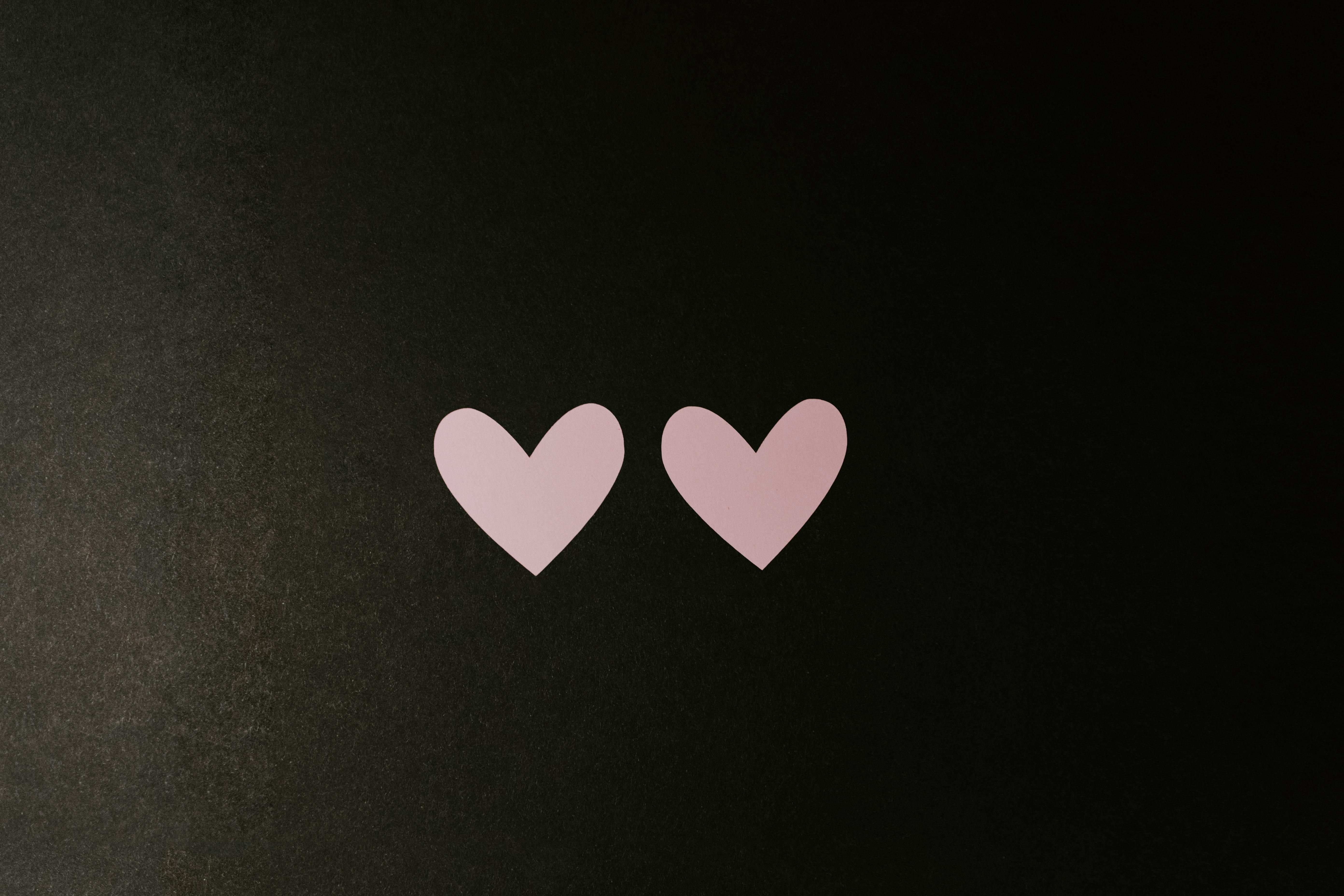 Két rózsaszín papírból kivágott szívecske egymás mellett egy sötét felületen