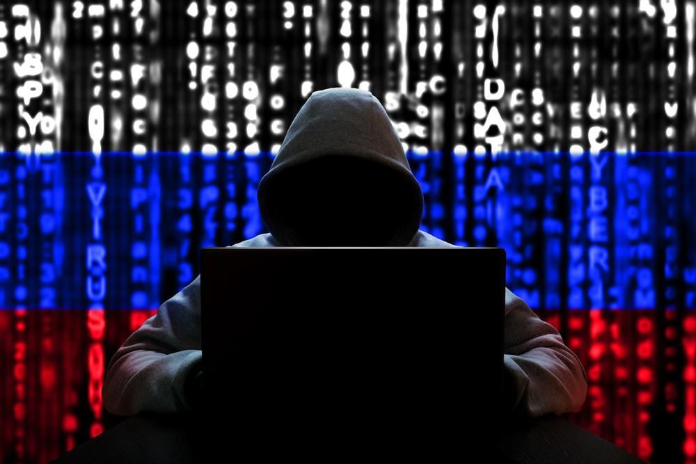 Kapucnis hacker ül a gépe előtt, a háttérben az orosz zászló látható