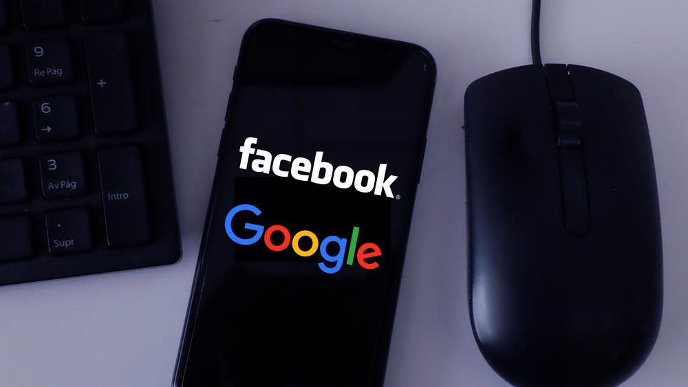 Egy fekete egér és klaviatúra között egy fekete iPhone van, aminek  a képernyőjén a Facebook és a Google logója látható