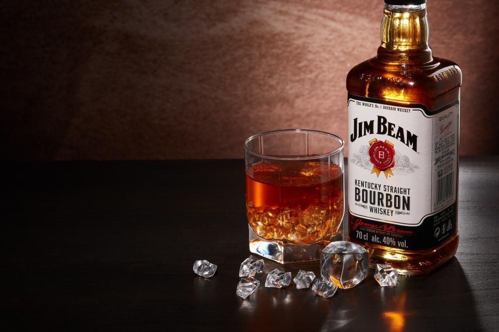Egy üveg Jim Beam whiskey egy pohár whisky mellett, előttük jégkockák láthatók