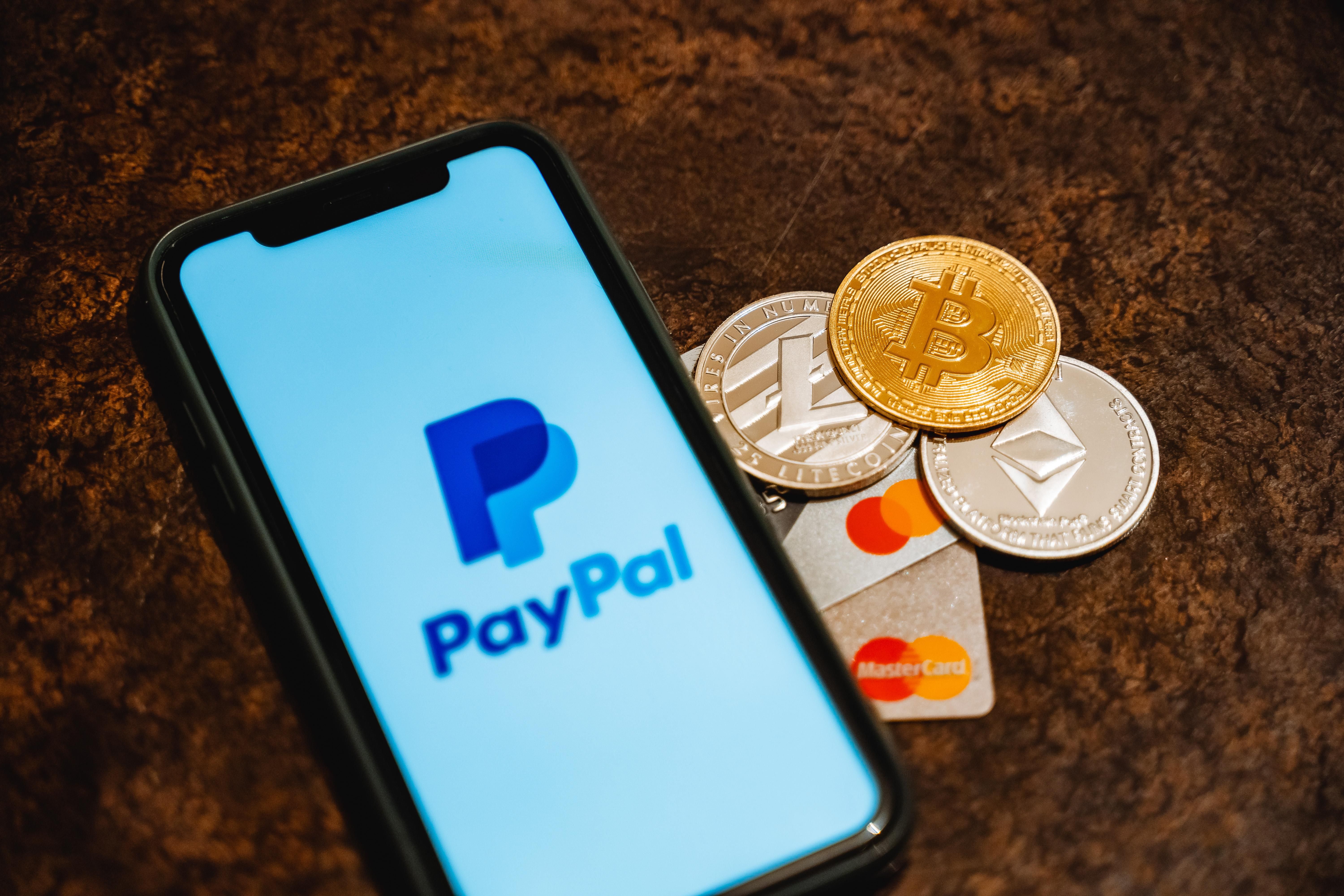 A PayPal logója egy okostelefon képernyőjén, mellette kriptovaluták és egy MasterCard bankkártya, a PayPal szankcionálja az oroszokat