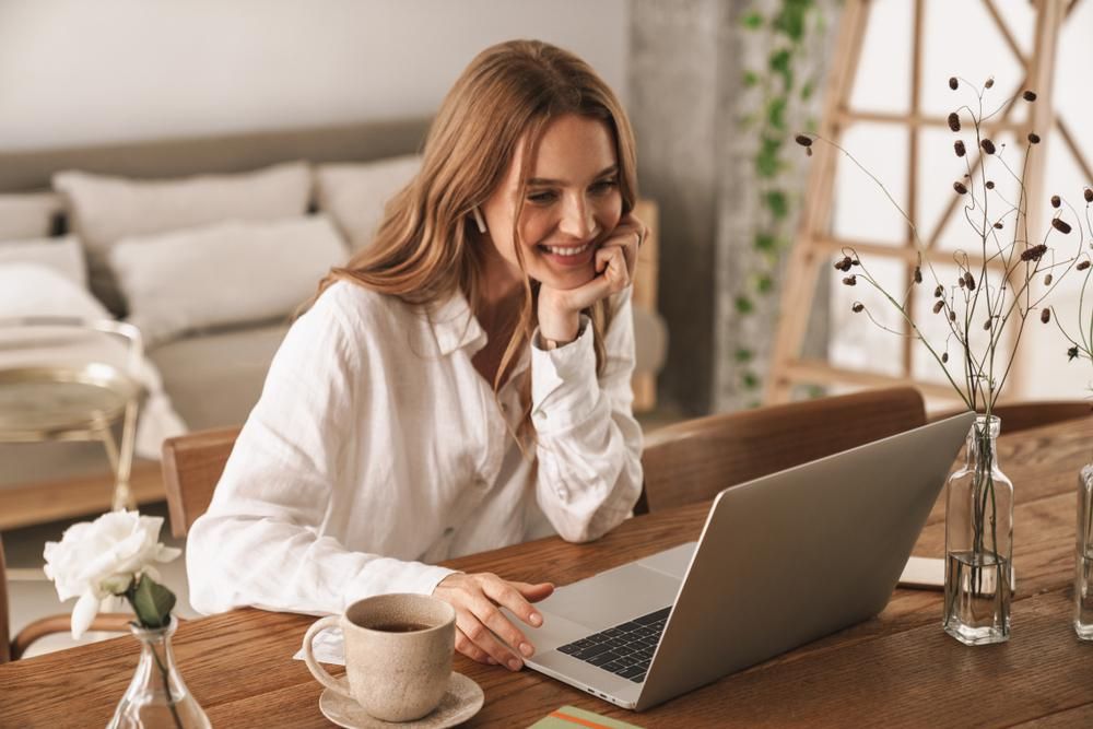 Szép lány fehér felsőben a laptopját nézi nevetve, home office-ból, otthonról dolgozik, munkaadója távmunka lehetőséget biztosít
