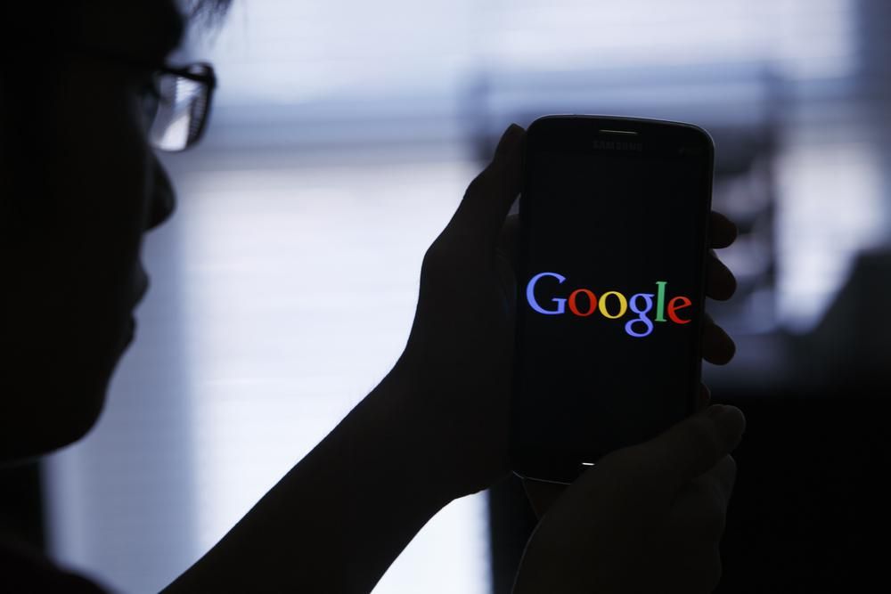 A gonosz pert megnyerő Google logója egy telefon képernyőjén, amelyet egy szemüveges ember tart a kezében, nagy a sötétség