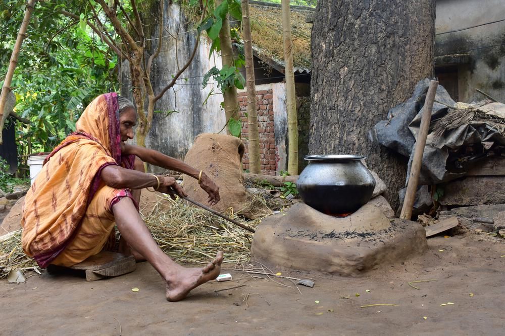 idős, szegény nő mezítláb egy bográcsban főz  a földön ülve