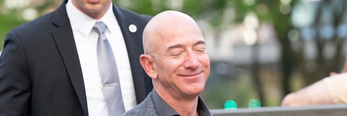 Jeff Bezos szürke öltönyben mosolyog egy fekete zakós ember előtt napsütéses időben, csukott szemmel