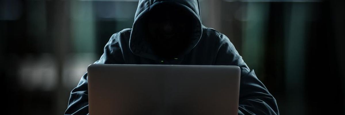 Kapucnis hacker ül a gépe előtt, ami egy fa asztalon van