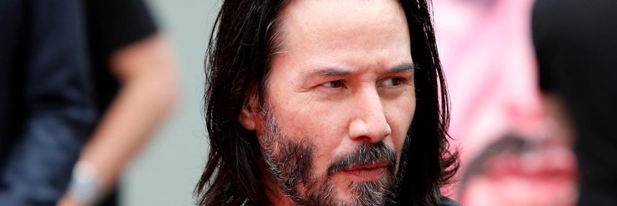 Keanu Reeves fekete öltönyben, hosszú hajjal, borostásan kémleli a környezetét