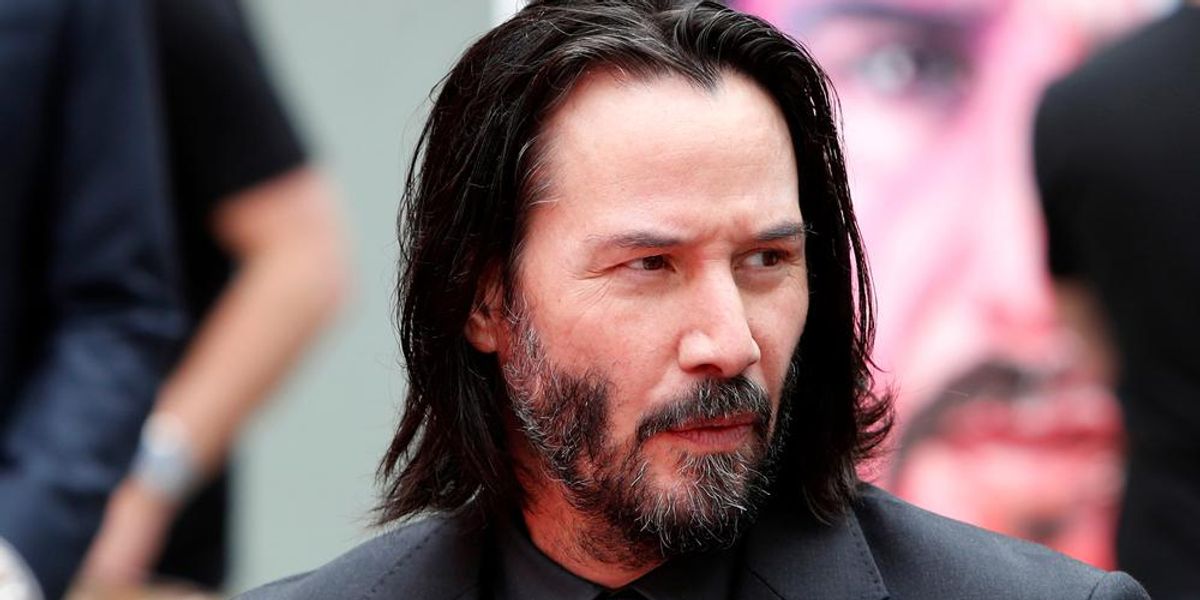 Keanu Reeves fekete öltönyben, hosszú hajjal, borostásan kémleli a környezetét