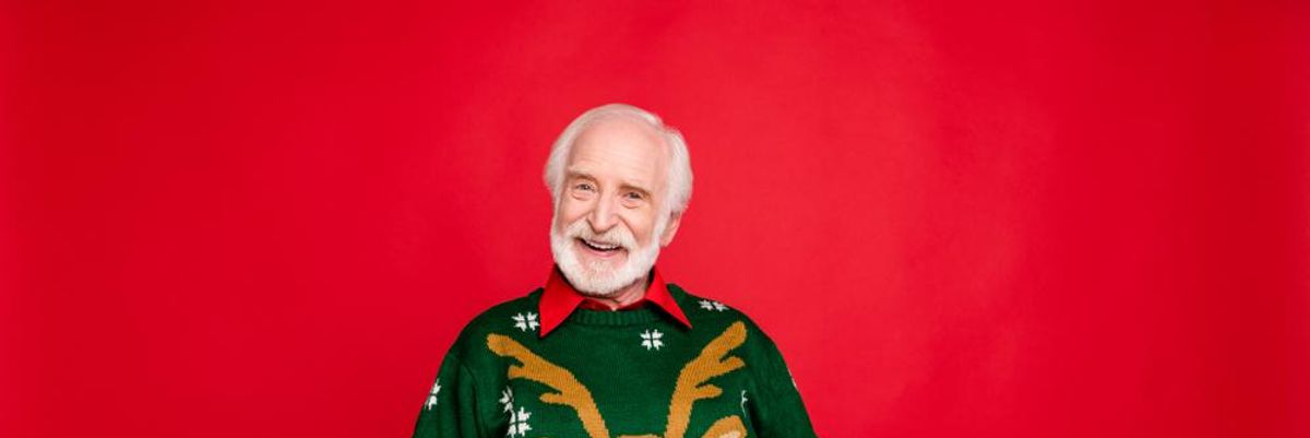 Kedves idős bácsi csúnya karácsonyi pulcsiban mosolyog egy piros fal előtt