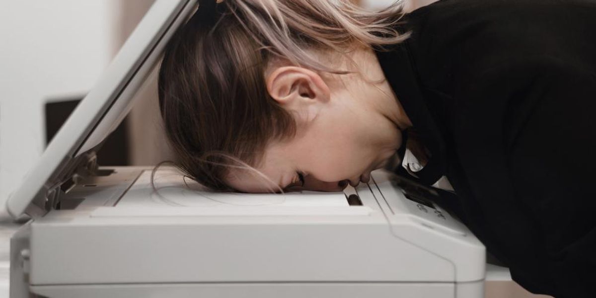 Kimerült szőkés-barna hajú nő fekete zakóban csukott szemmel beletette a fejét a munkahelyi nyomtatóba