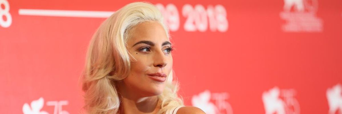 Lady Gaga fehér ruhában, szőke hajjal, mosolyogva tündököl egy díjátadó gálán egy piros-fehér molinó előtt