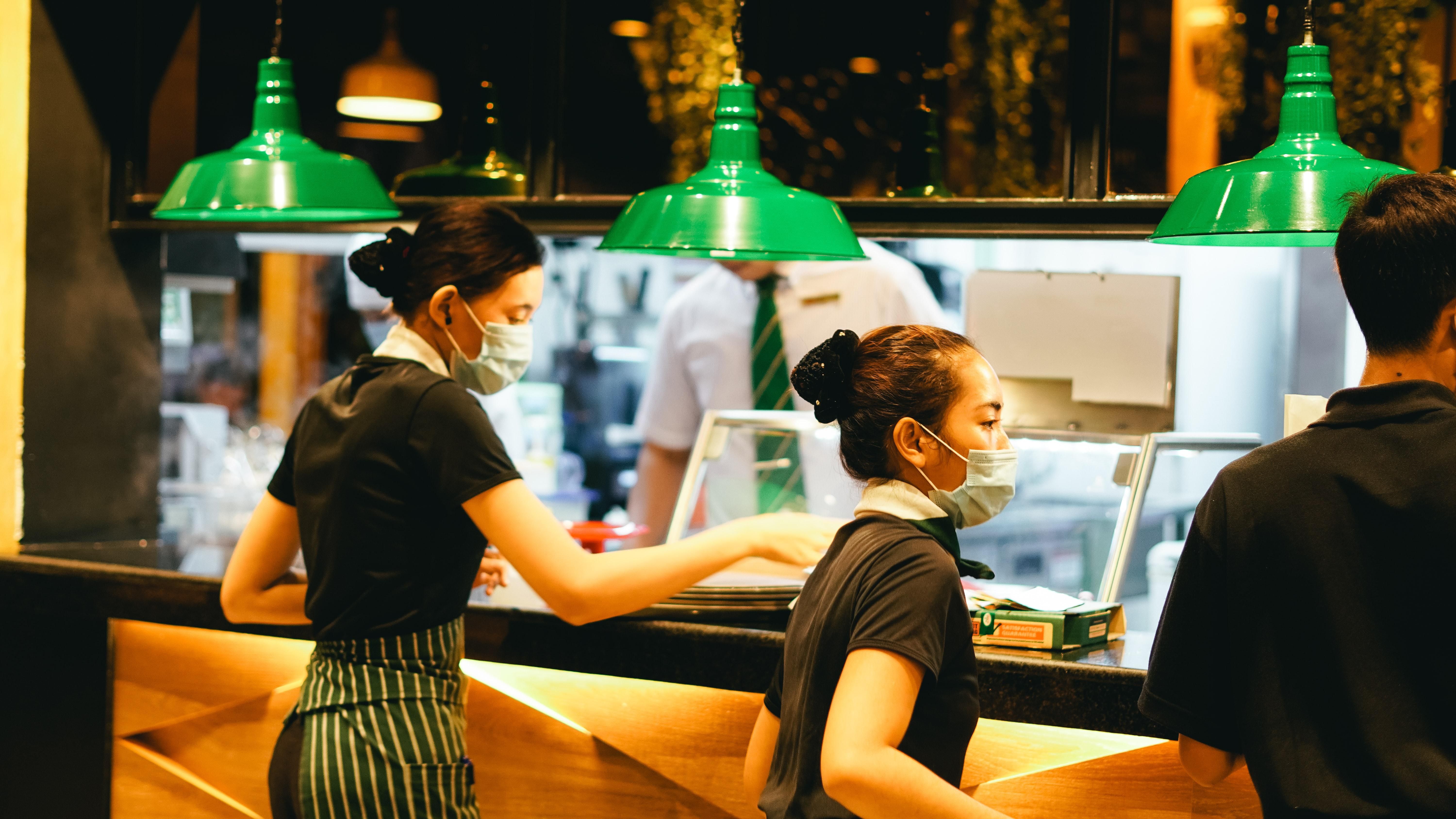 Arcmaszkos, fekete felsős, zöld-fehér csíkos kötényes pincérnők szolgálják fel az ételeket a vendégeknek egy étteremben