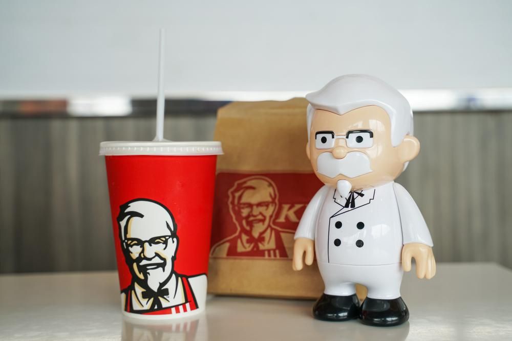 Egy KFC-s üdítő és egy papírzacskó mellett Sanders ezredes babája látható egy fehér asztalaon