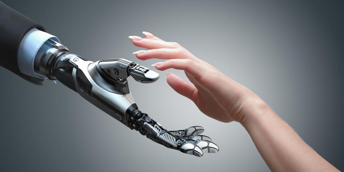 Mesterséges intelligenciával felvértezett robot nyújtja a kezét egy kifestett körmű nőnek egy szürke szobában