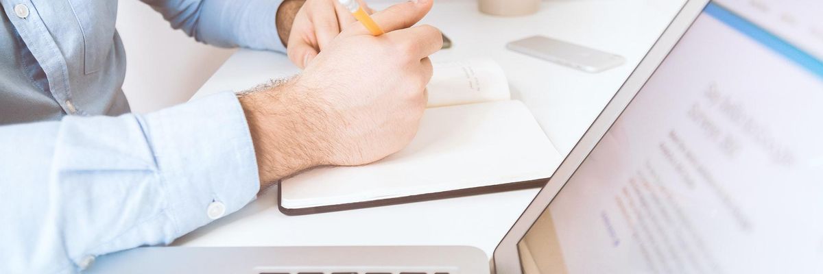 Nyitott ezüst laptop kéz ír egy noteszba íróasztalnál