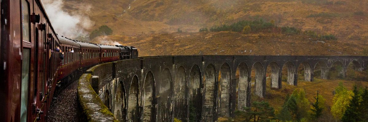 Piros vonat halad át egy hídon dombok között füstölve