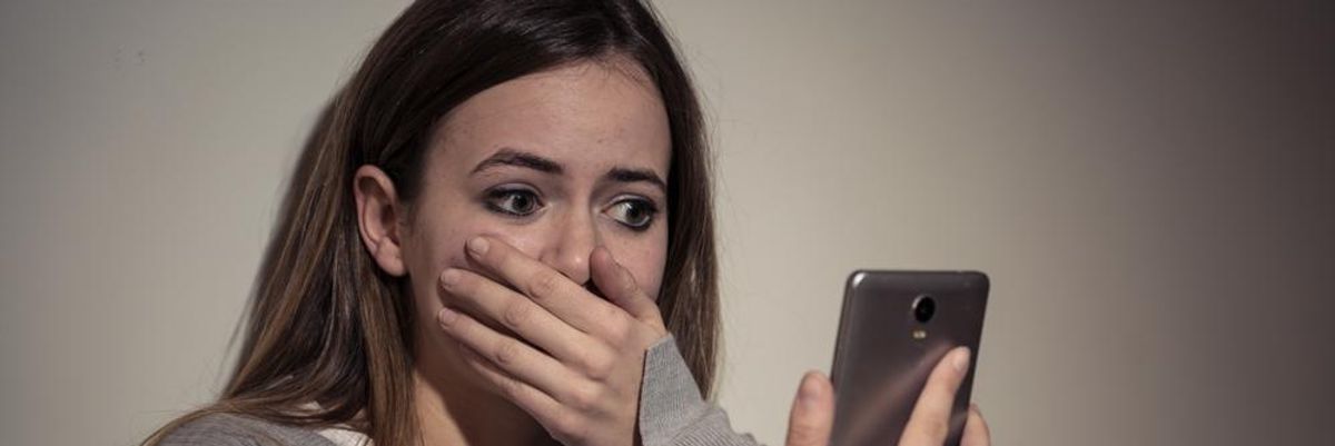 Szomorú fiatal tinédzser lány elképedve nézi a telefonján a Facebook-on megjelenő felzaklató posztokat fehér pólóban, szürke pulcsiban