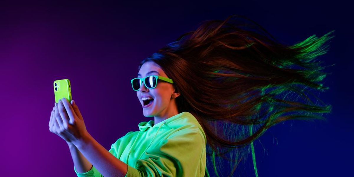 Zöld pulcsis lánynak lobok a haja az internet gyorsaságától miközben a zöld napszemüvegén keresztül nézi a sárga telefontokos okostelefonját egy lilás-kékes háttérben
